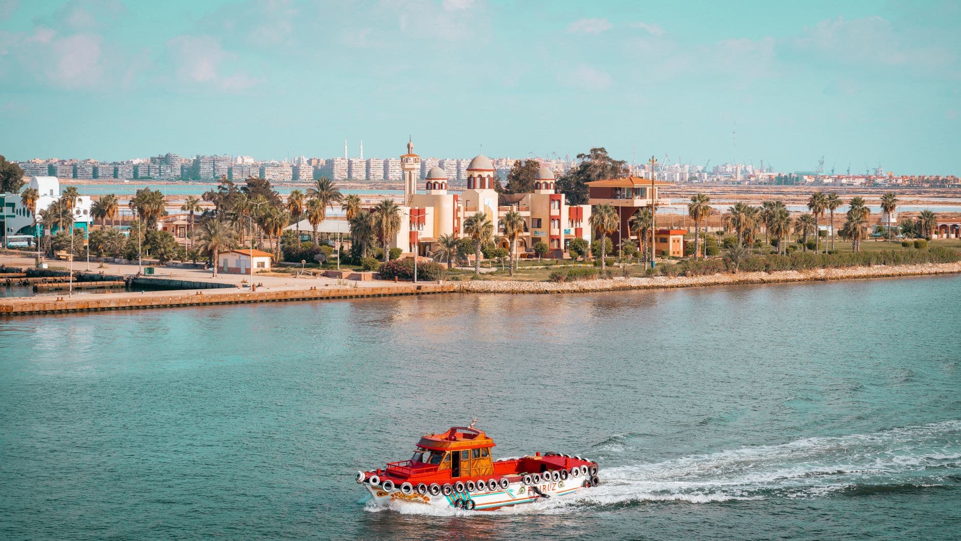 A scenic view of a red boat cruising near a city with palm trees and vibrant architecture.
