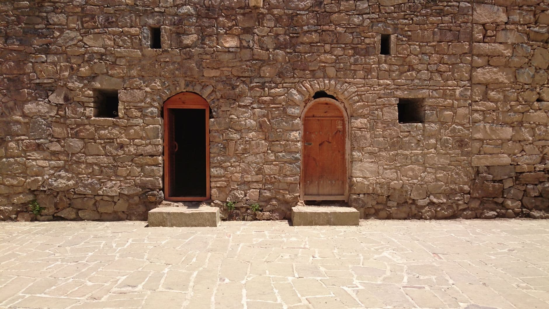 An ancient stone building facade with two wooden arched doors under bright sunlight.