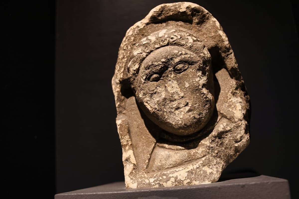 A weathered stone bust of a person with a head covering on a pedestal.