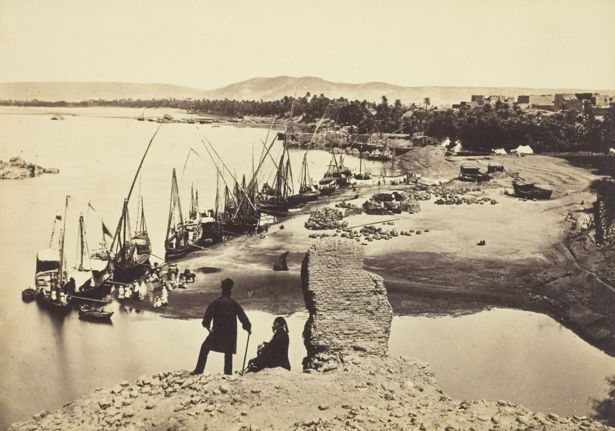 Sailboats line a riverbank with distant buildings and hills.