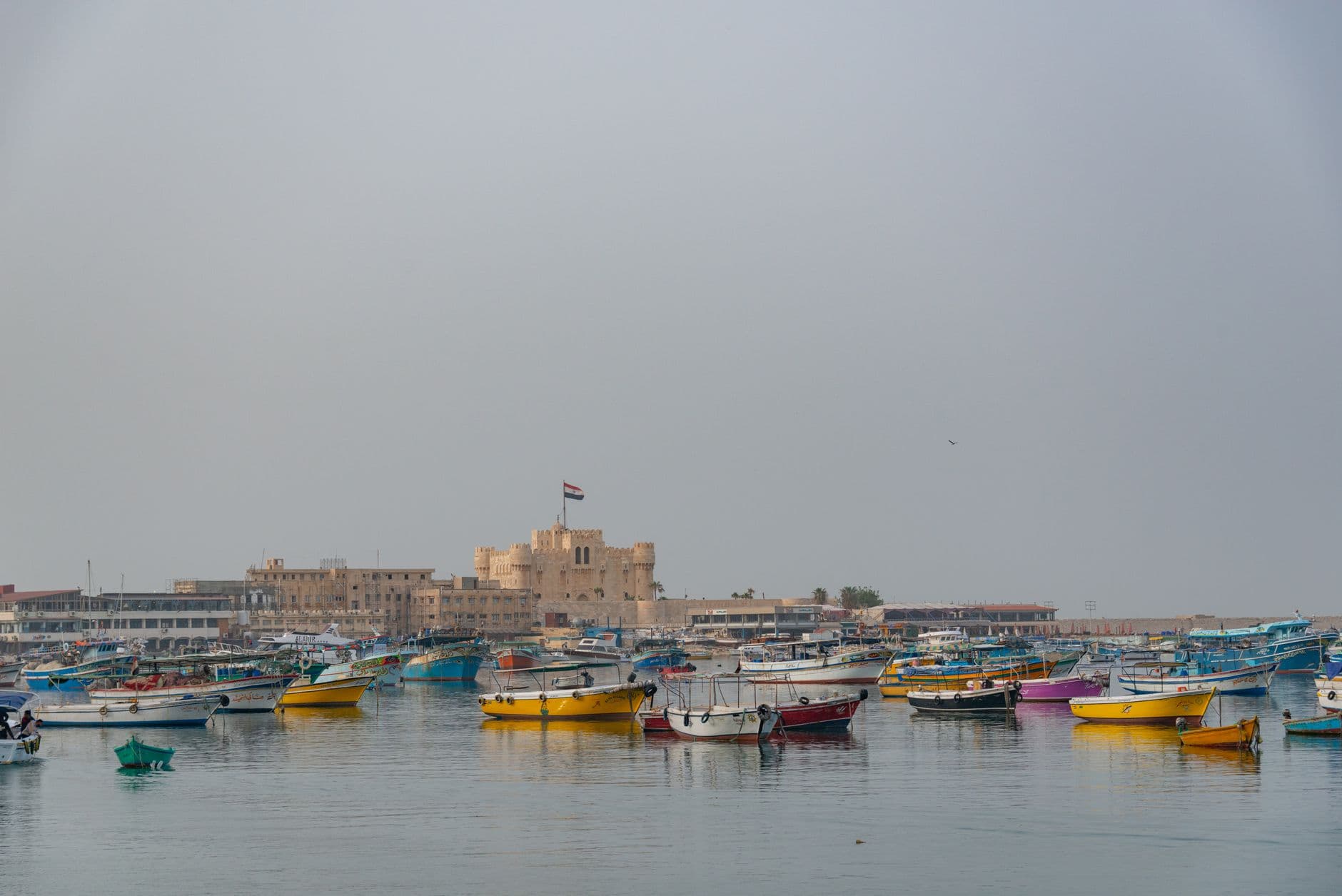 Colorful boats float in Alexandria's harbor with the historic Citadel of Qaitbay in the background.